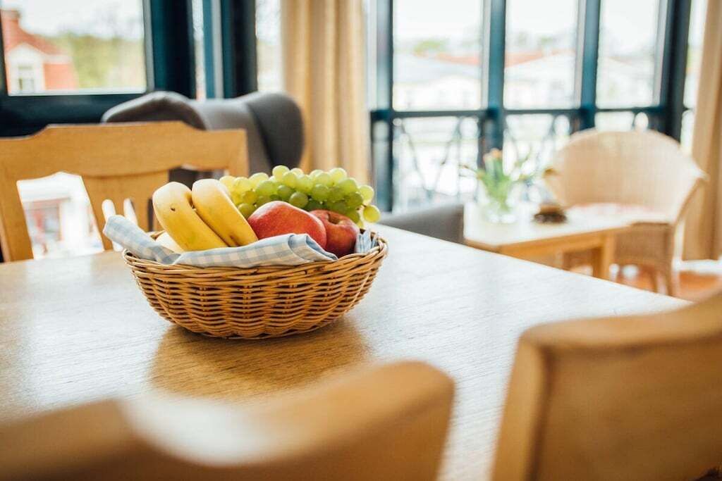 A wicker fruit basket on a wooden table with a view of a cityscape through large windows.