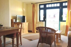 A room with a television on a stand, a dining table with chairs, a small table with a chair, and a window with curtains. - Tower House Holiday Apartment 11 (Kühlungsborn)