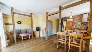 Uma sala de estar ampla com piso de madeira, um sofá, uma mesa de jantar com cadeiras e uma cozinha com parede de tijolos e vigas de madeira.