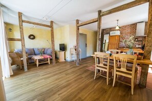 Amplia sala de estar con piso de madera, un sofá, una mesa de comedor con sillas y una cocina con pared de ladrillo y vigas de madera.