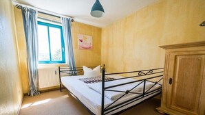Uma cama de solteiro com estrutura de metal, um guarda-roupa de madeira, um radiador e uma janela com cortinas.