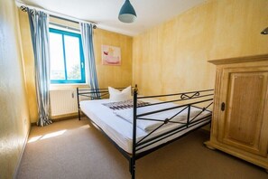 Una cama individual con estructura metálica, un armario de madera, un radiador y una ventana con cortinas.