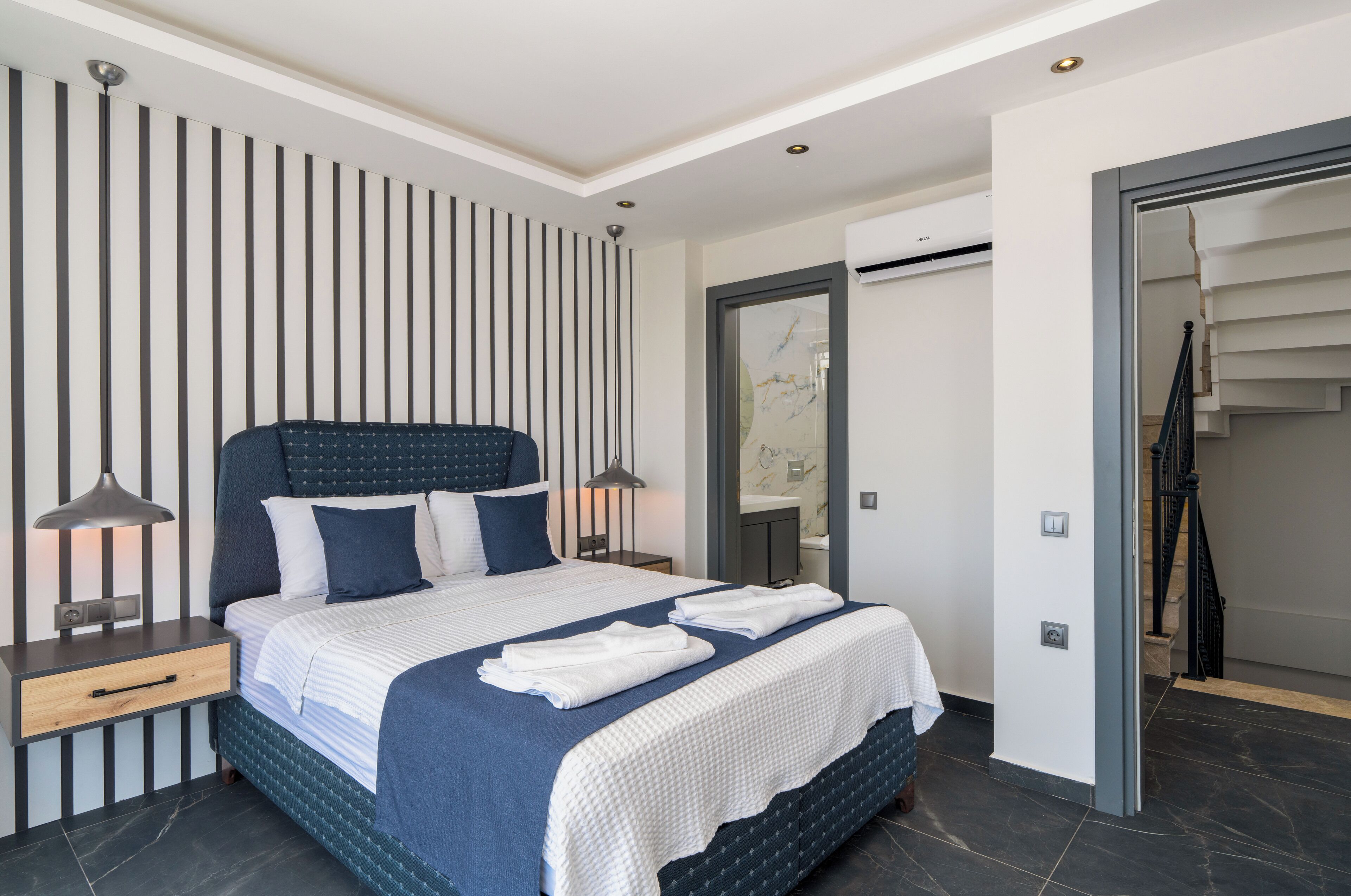 Un dormitorio moderno con cama, lámparas de noche, mesita de noche y un baño visible a través de una puerta abierta.