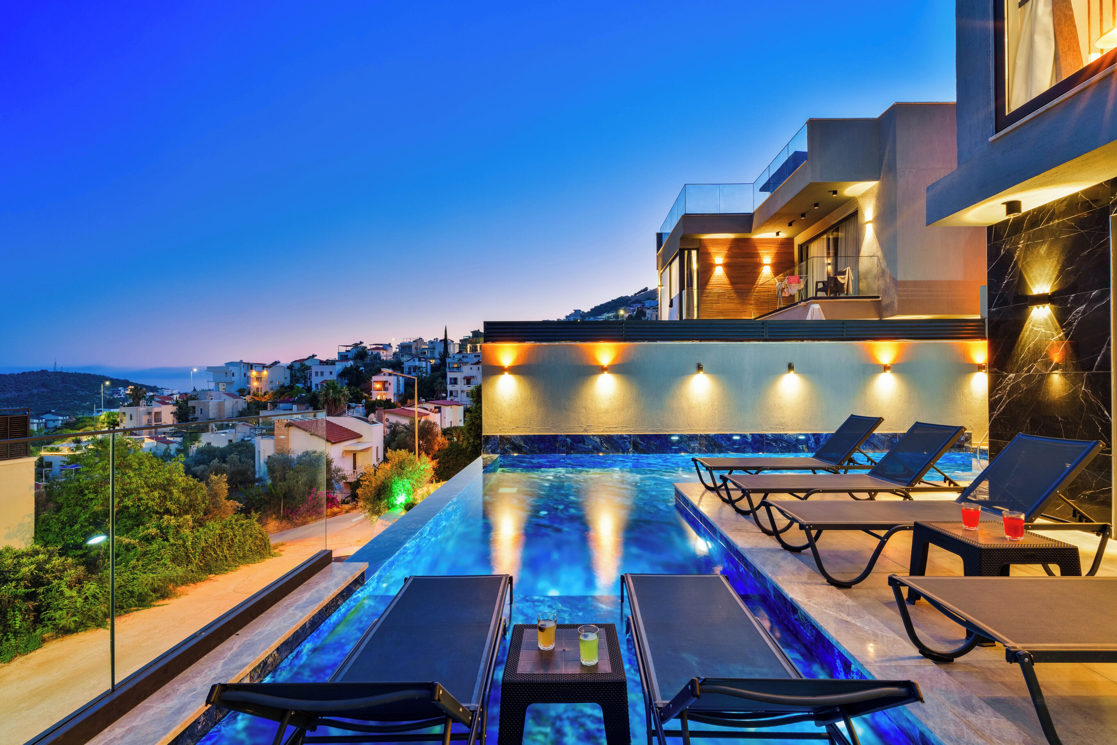 A rooftop infinity pool with lounge chairs and a table, overlooking a cityscape at dusk.