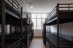 Un dormitorio con literas dispuestas en fila, una ventana con vista a árboles desnudos y un radiador en el suelo.