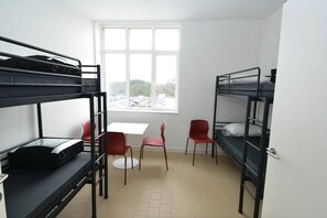 A dormitory room with bunk beds, a table, chairs, and a window with a view of parked cars.
