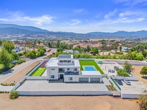A modern two-story house with a flat roof, a tennis court, and a parking area. - Villa Spectacular - Luxury Hilltop Estate | Wine Country Views (Temecula)