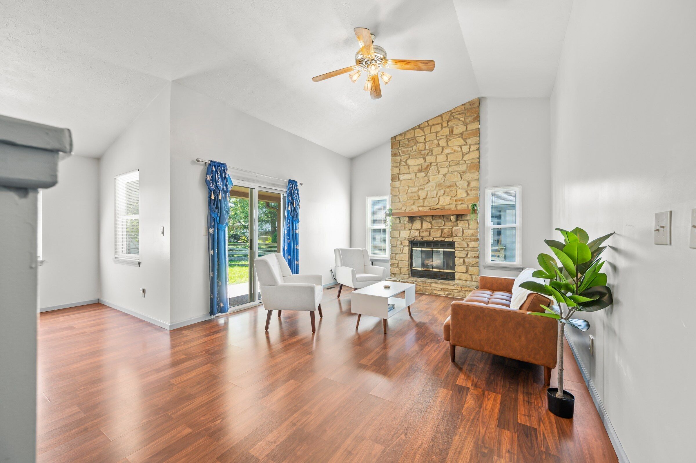 A spacious living room with hardwood flooring, a stone fireplace, and a ceiling fan.