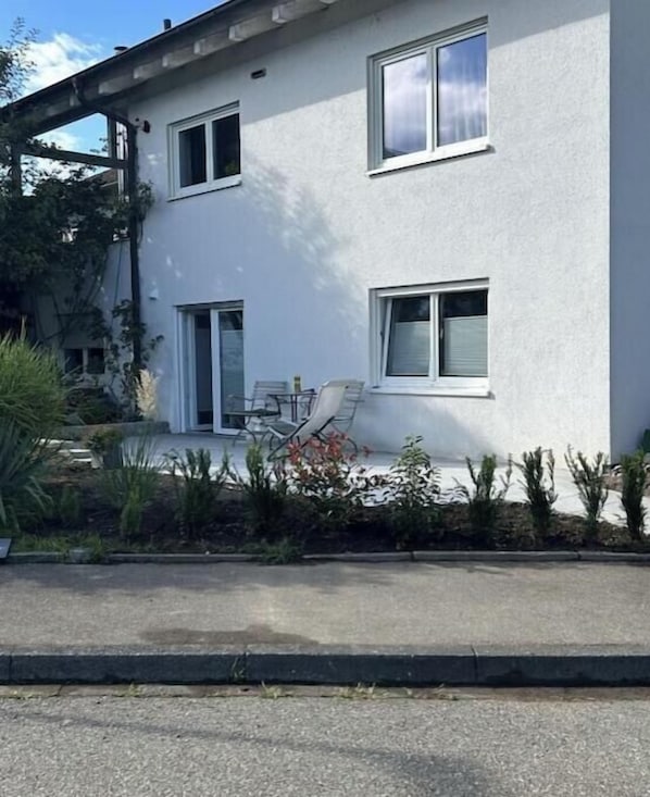 Un bâtiment à deux étages, à l’extérieur blanc, doté d’un patio couvert avec des meubles d’extérieur et d’un petit jardin agrémenté de diverses plantes.