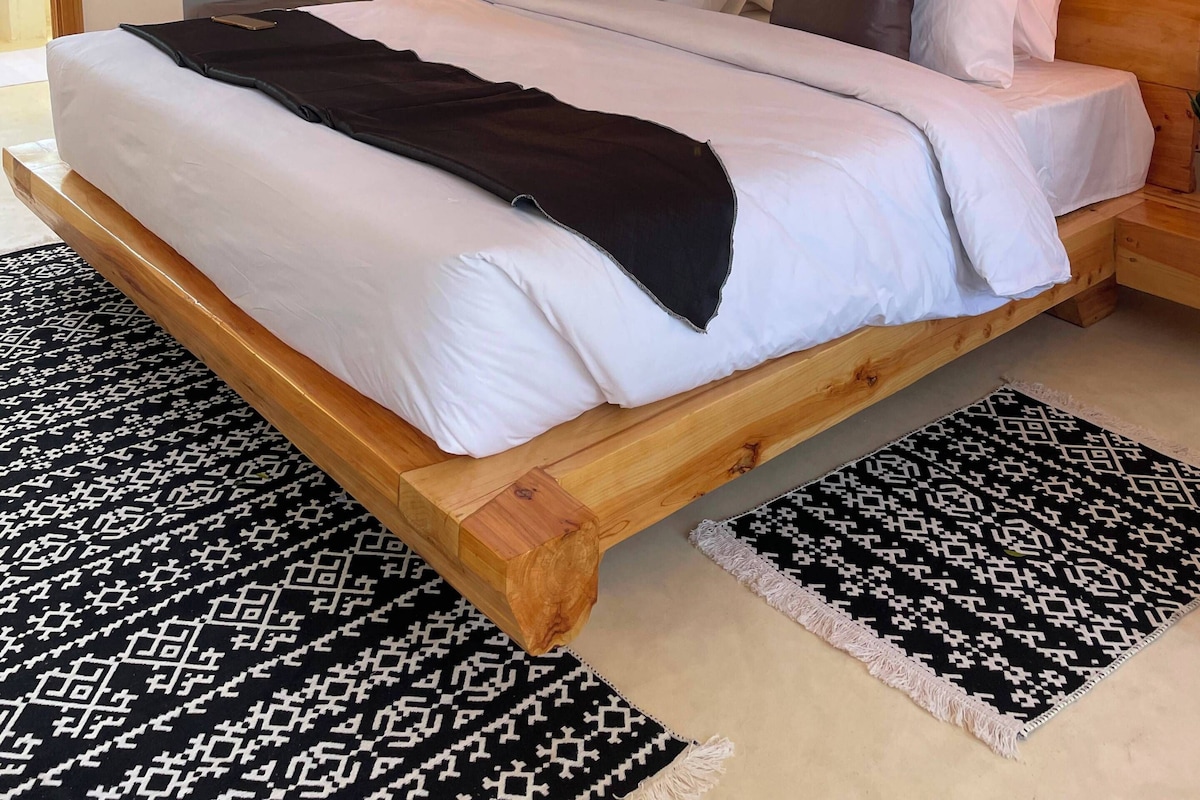 Cama com estrutura de madeira, roupa de cama branca e manta preta.