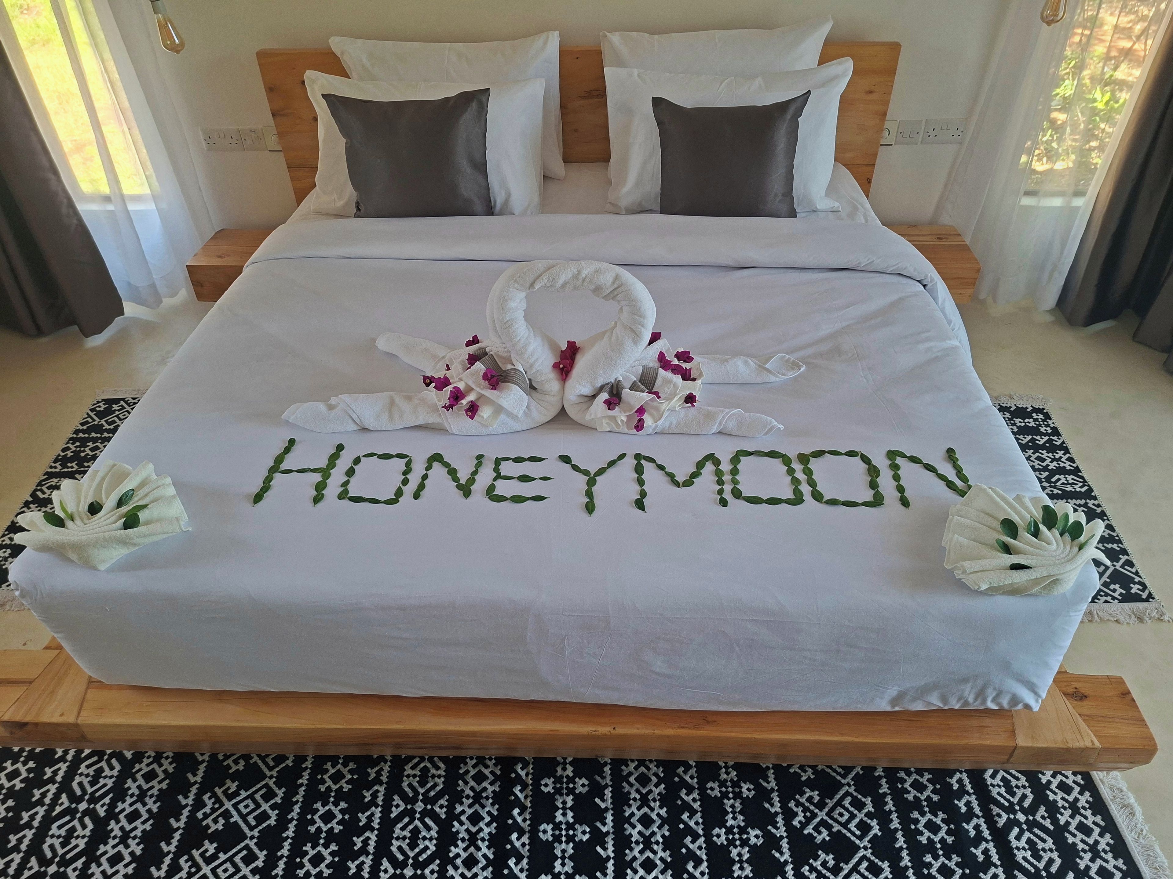 Una cama impecablemente arreglada con una toalla y flores dispuestas para deletrear 'HONEYMOON' sobre el edredón.