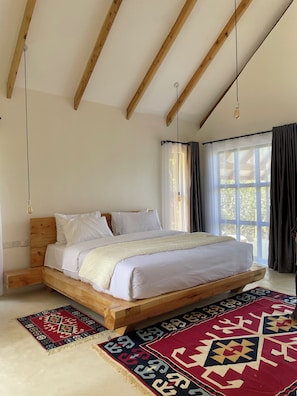 Quarto com estrutura de cama de madeira, roupa de cama branca, luminária pendente e uma janela grande com cortinas.