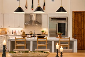 A modern kitchen with white cabinetry, a central island with bar stools, a stainless steel range hood, and a large refrigerator.