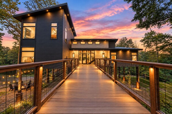 A modern two-story house with a wooden deck leading to the entrance, surrounded by trees.