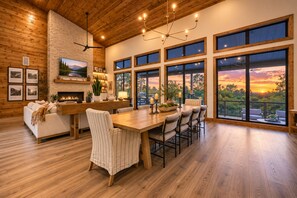 A spacious dining area with a large wooden table and chairs, a fireplace, and large windows offering a view of the sunset.