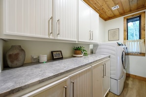 A kitchen with white cabinets, a marble countertop, a washing machine, and a window with a view of trees.
