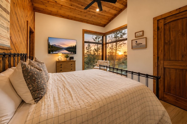 A bedroom with a large bed, a nightstand, a lamp, a painting, and a view of the outdoors.