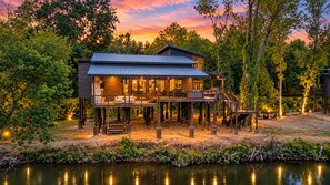 A two-story wooden house with a deck, situated by a body of water, surrounded by trees.