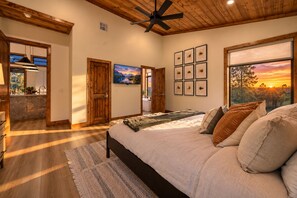 A bedroom with a bed, a ceiling fan, a large window with a sunset view, and a painting on the wall.