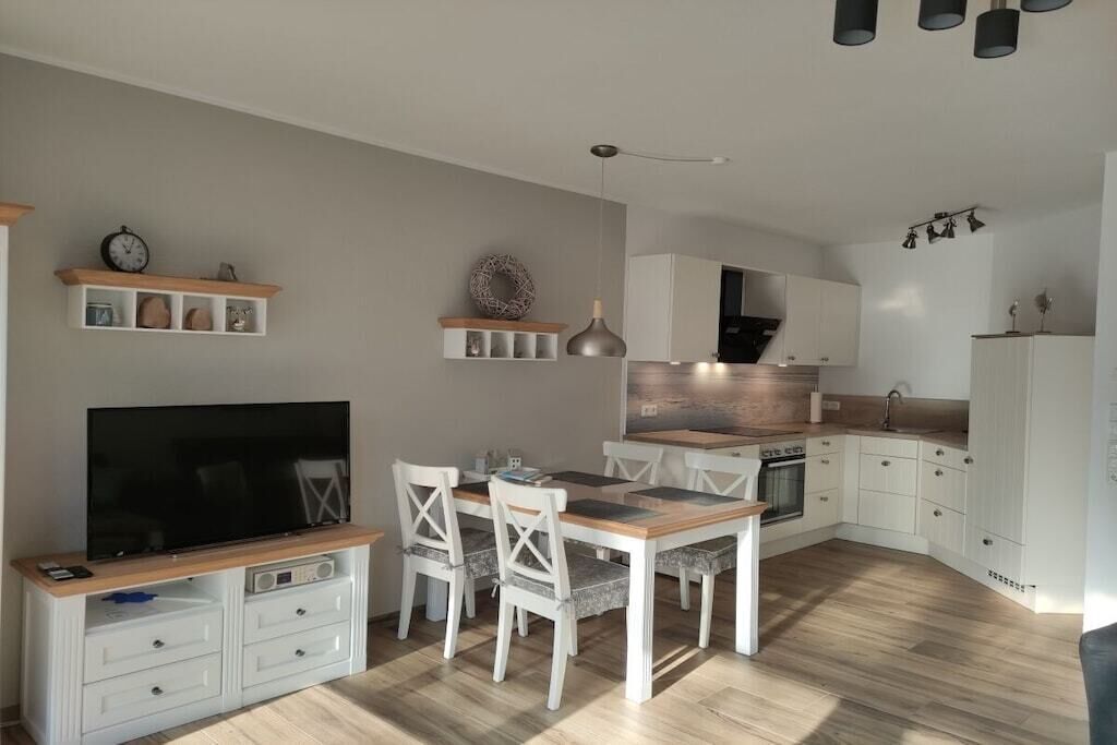 A modern kitchen with white cabinets, a dining table with chairs, a flat-screen TV, and a pendant light.