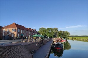 A canal-side walkway with a brick building, a row of tables and chairs, and a boat docked on the water. - Holiday home with 1 bedroom (Wangerland)