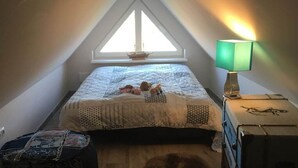 Quarto com teto inclinado, cama com colcha, mesa de cabeceira com abajur e janela com vista.