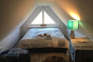 A bedroom with a sloped ceiling, a bed with a quilt, a nightstand with a lamp, and a window with a view. - Holiday home with 3 bedrooms (Nienhagen)