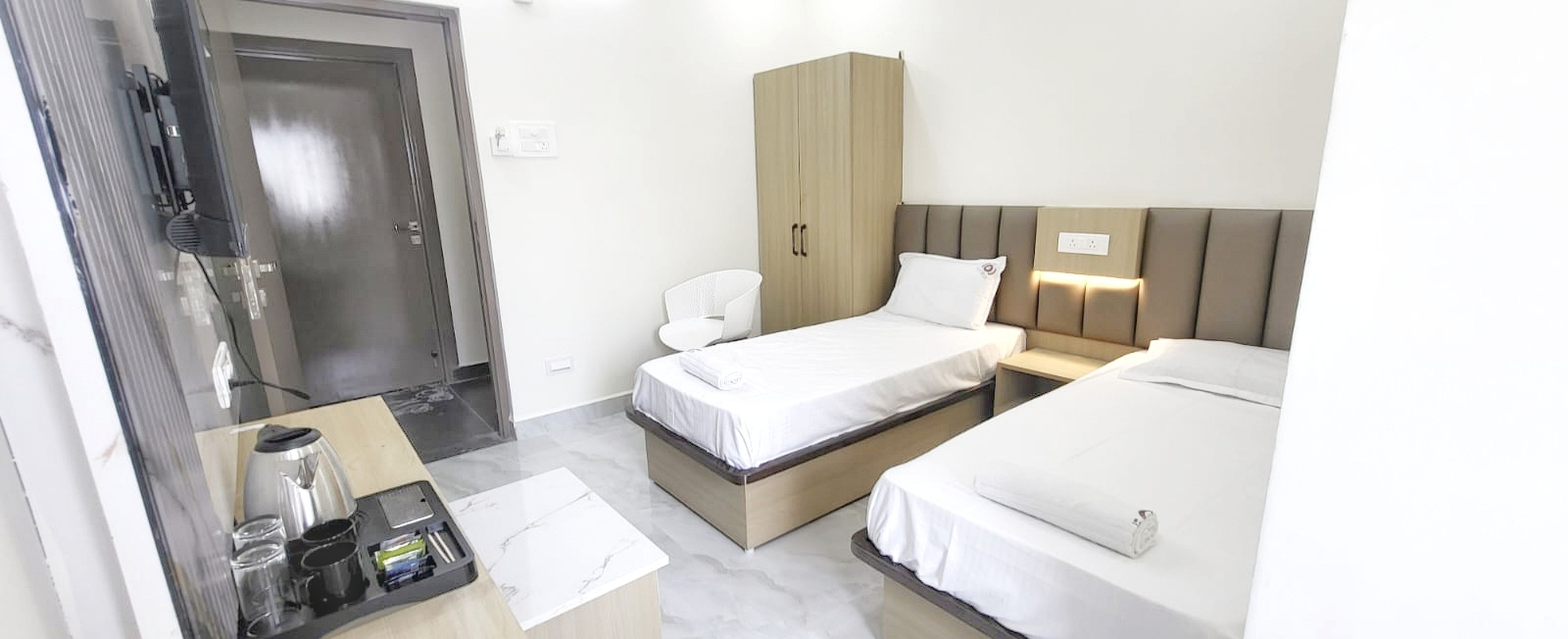 A hotel room with a bed, a wardrobe, a bedside table with a kettle, and a chair.