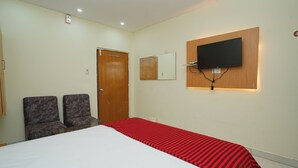 Ein Hotelzimmer mit einem Bett, zwei Stühlen, einer Holztür, einem an der Wand befestigten Fernseher und einem Wandspiegel.