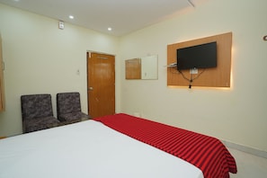 Een hotelkamer met een bed, twee stoelen, een houten deur, een wandtelevisie en een spiegel aan de muur.