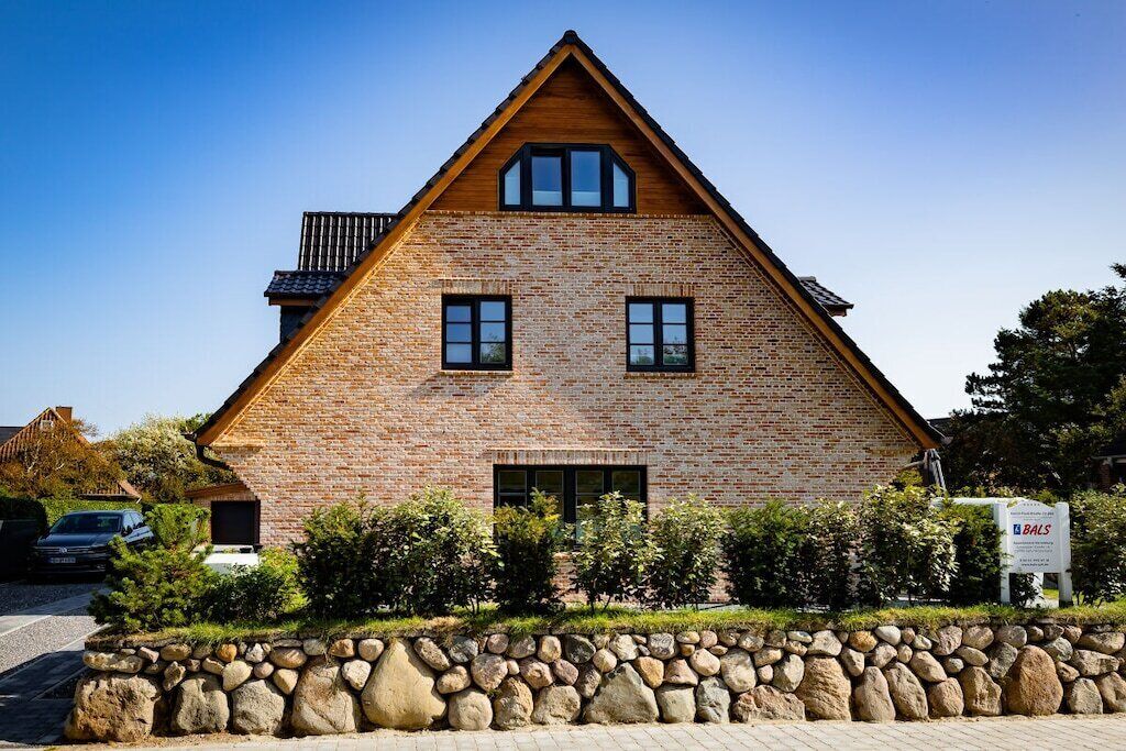 Una casa de ladrillo con techo triangular, dos ventanas y un muro de piedra frontal.