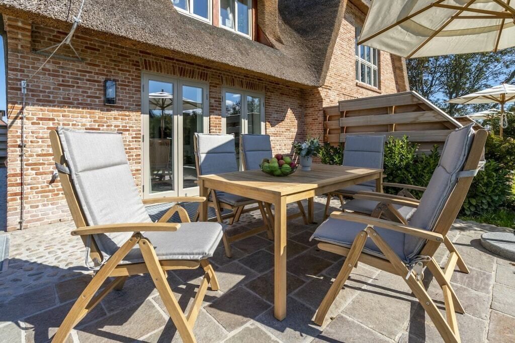 An outdoor patio with a wooden table and chairs, a thatched roof, and a brick house in the background.