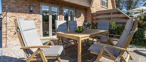 Um pátio externo com mesa e cadeiras de madeira, um telhado de palha e uma casa de tijolos ao fundo.