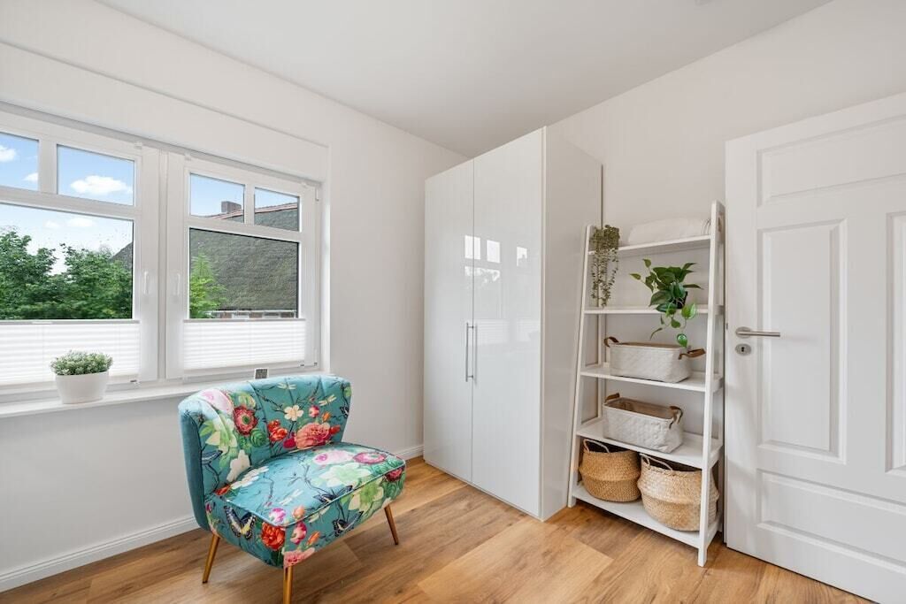 Ein Zimmer mit Holzboden, einem blumigen Sessel, einem weißen Kleiderschrank und einer weißen Tür.