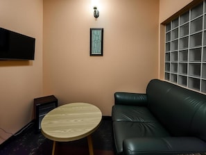 Een woonkamer met een zwarte leren bank, een ronde houten tafel, een lijst met abstracte kunst aan de muur, en een televisie boven de bank.