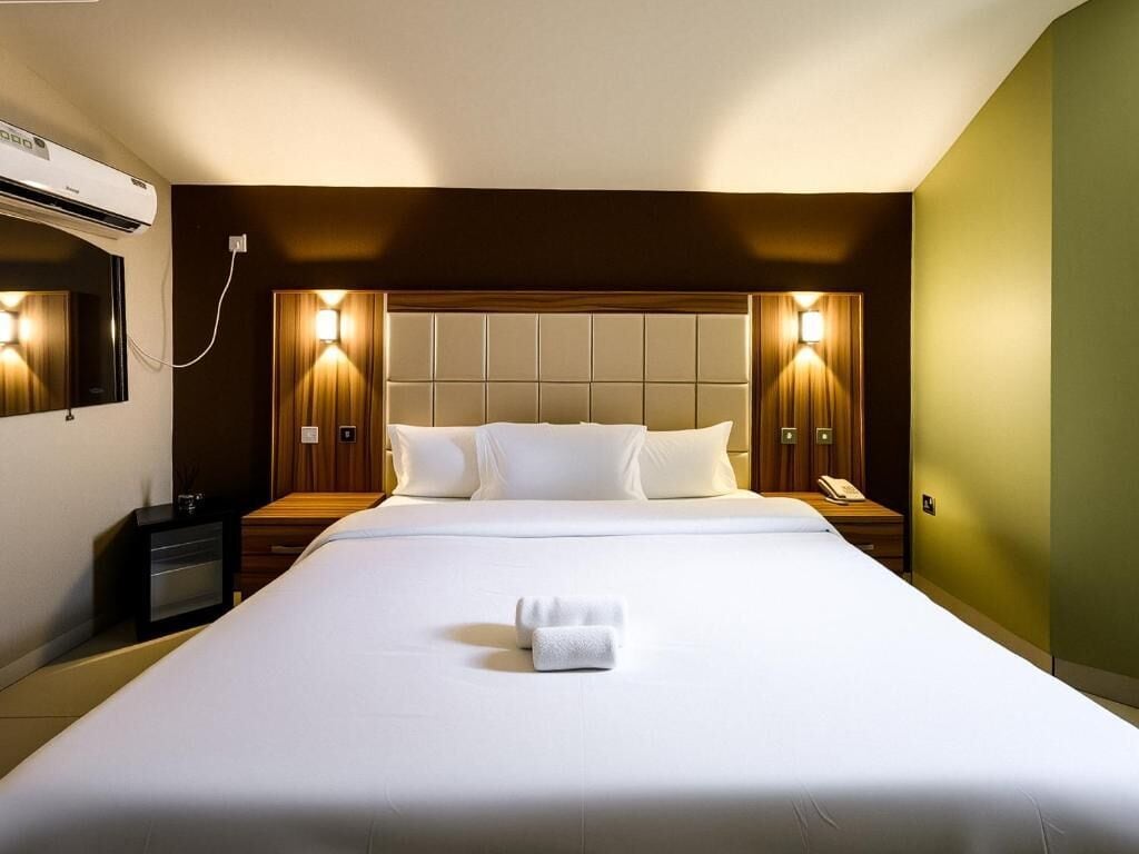 A hotel room with a large bed, two bedside tables, a nightstand, a wall-mounted air conditioner, and two wall sconces.
