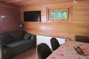 A room with a wooden wall, a flat-screen TV mounted above a window, a sofa, a dining table with chairs, and a radiator below. - Holiday home Berghaus (Reinhardshagen)