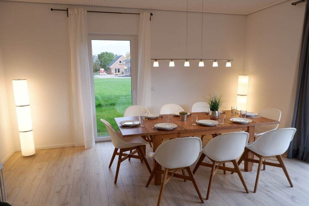 A dining area with a wooden table and white chairs, a potted plant, and a view of a house through a glass door.