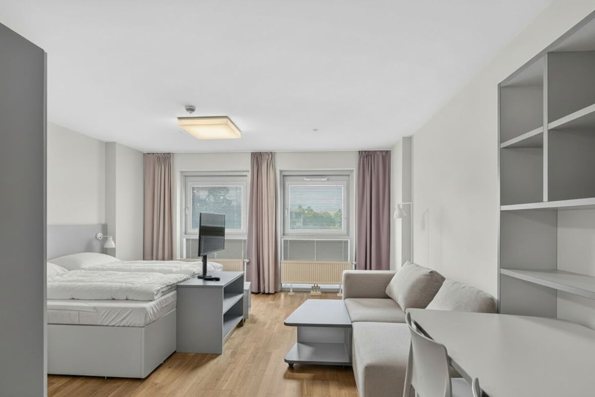 Ein modernes Hotelzimmer mit Bett, Schreibtisch, Sofa und einem großen Fenster mit Vorhängen.