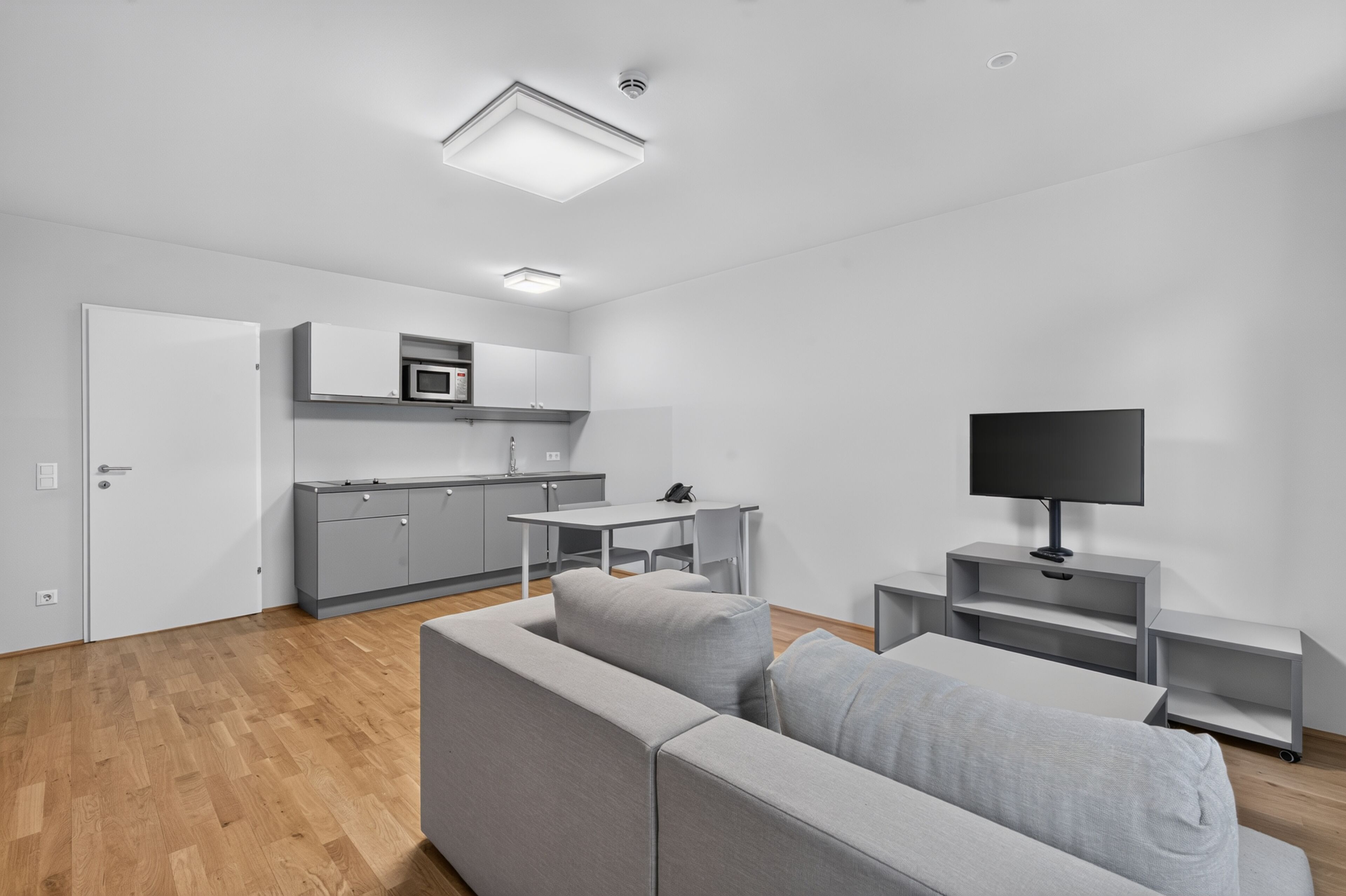 A modern living room with a grey sofa, a television on a stand, and a kitchen area with grey cabinets and a microwave.
