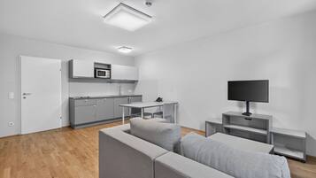 A modern living room with a grey sofa, a television on a stand, and a kitchen area with grey cabinets and a microwave.