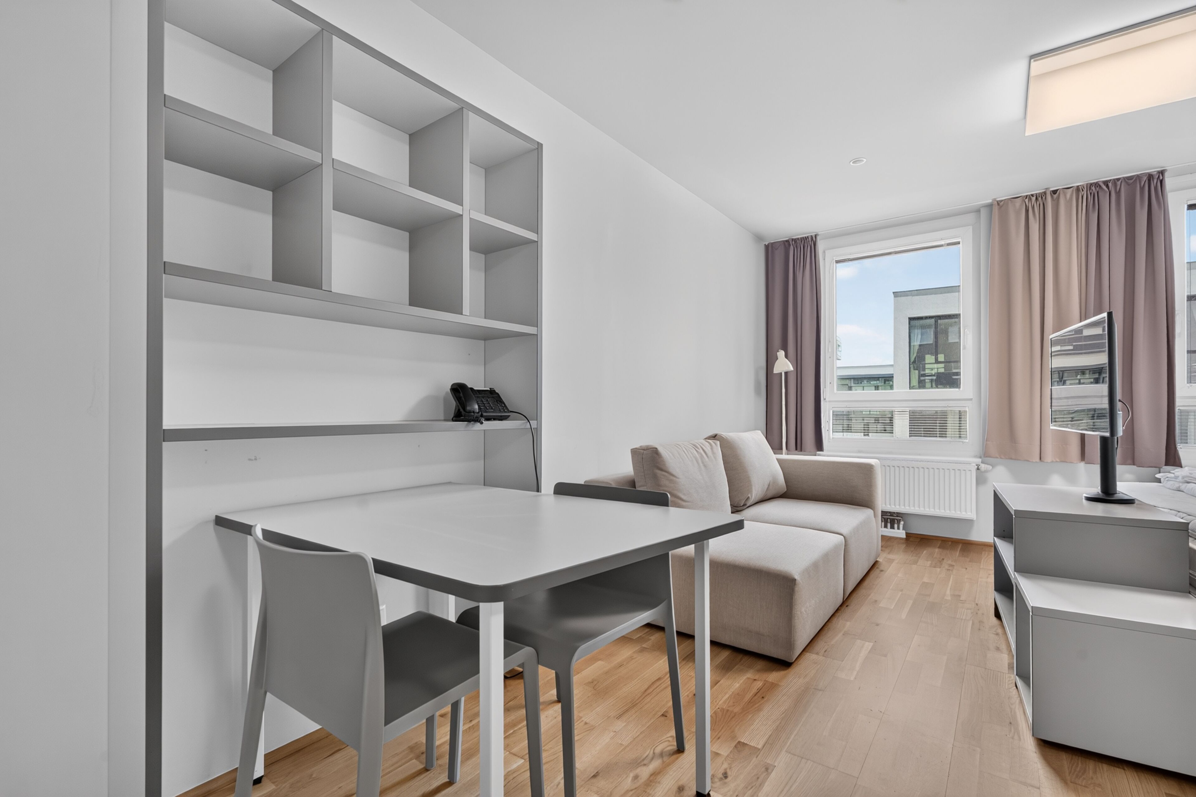 A modern, minimalist room with a dining table, two chairs, a sofa, and a small kitchenette area.