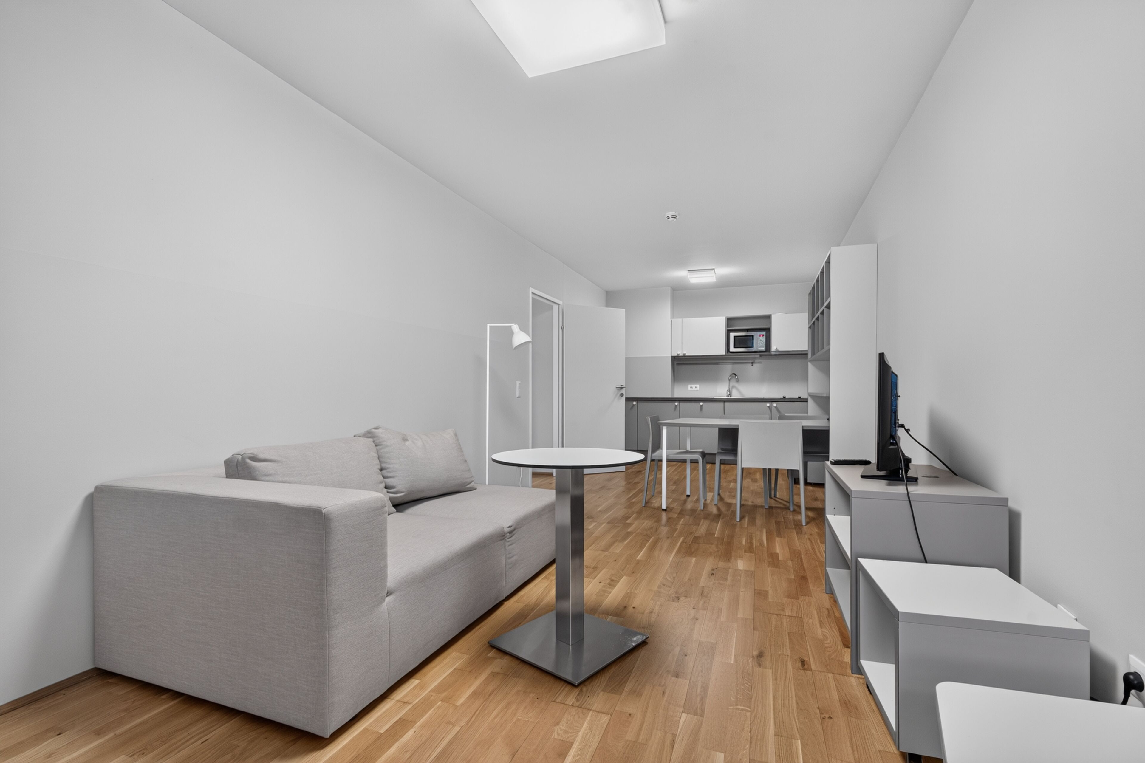 Un espacio habitable moderno y minimalista con un sofá gris, una mesa redonda y una cocina al fondo.