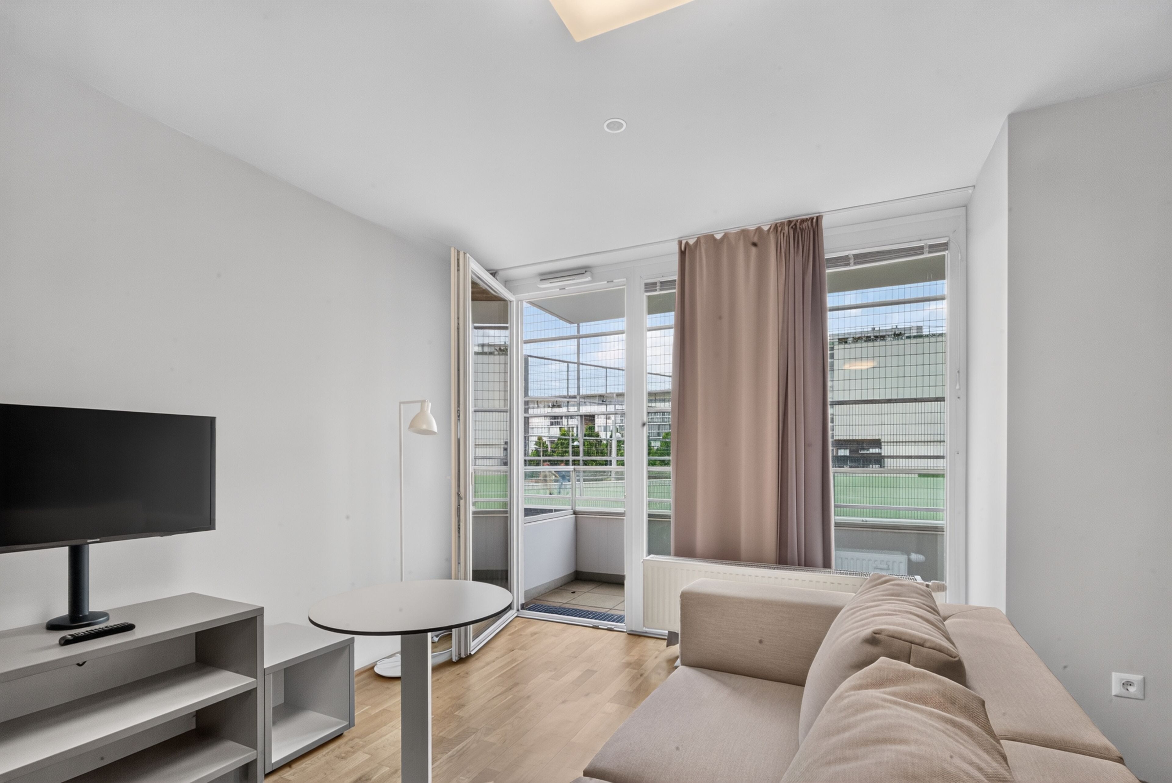 A modern living room with a flat-screen TV, a round table, a sofa, and a large window with curtains.