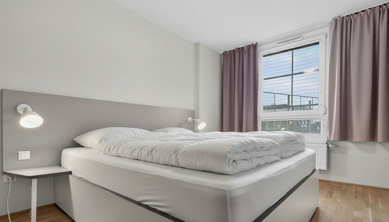 A modern bedroom with a bed, bedside lamps, a window with curtains, and a radiator.