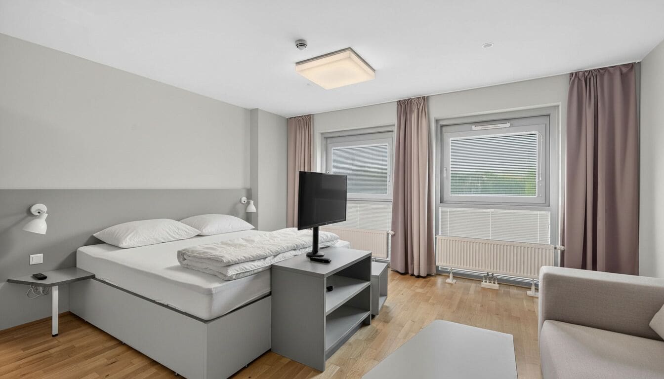 A modern hotel room with a bed, bedside table, TV, and a seating area.