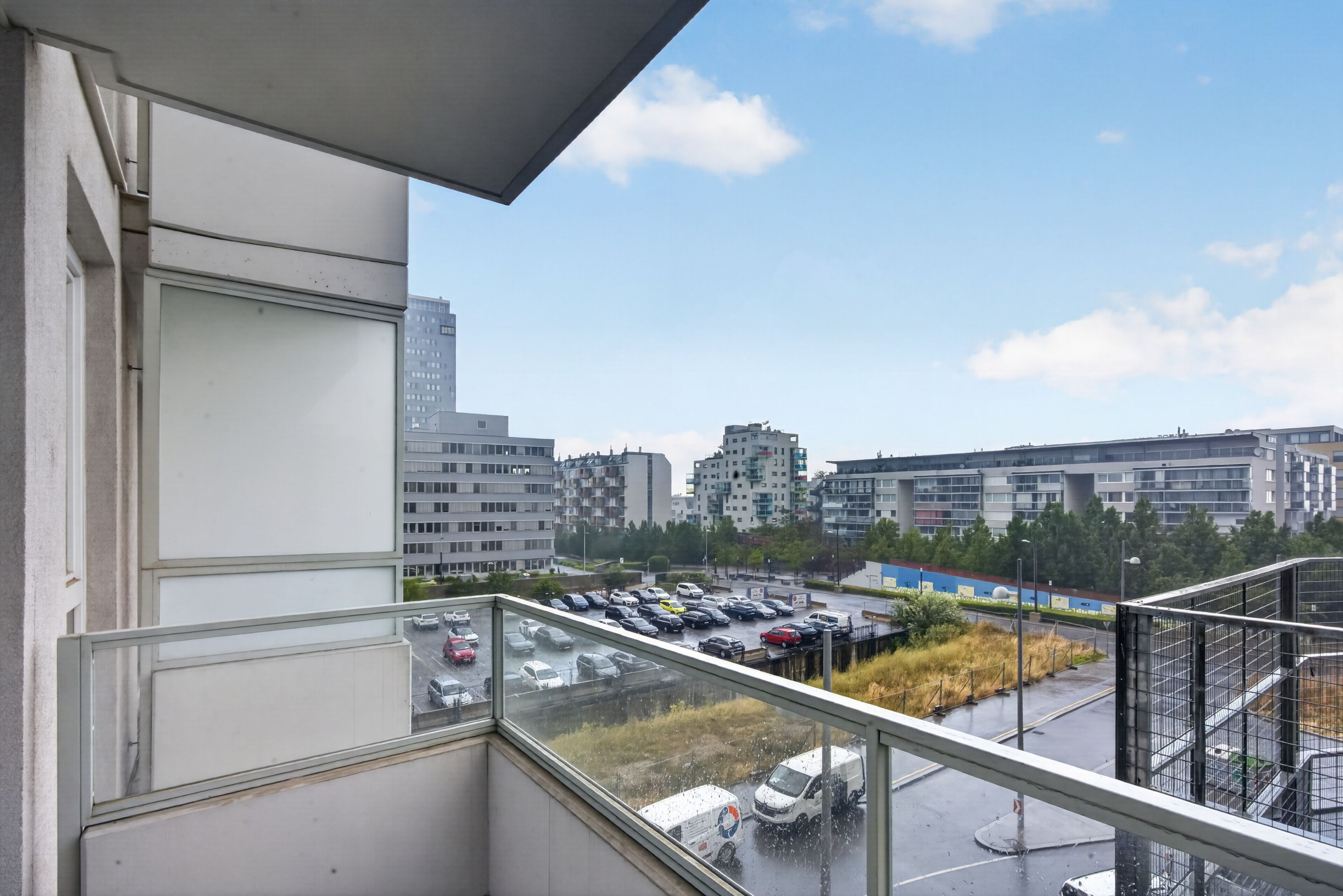 A view from a balcony overlooking a parking lot with multiple cars and a building in the background.