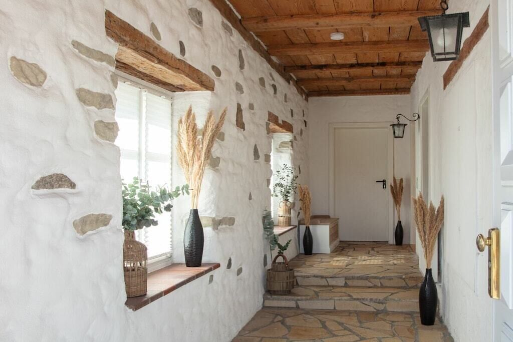 Passadís de pedra amb bigues de fusta rústica, una porta blanca i gerros decoratius amb plantes seques.