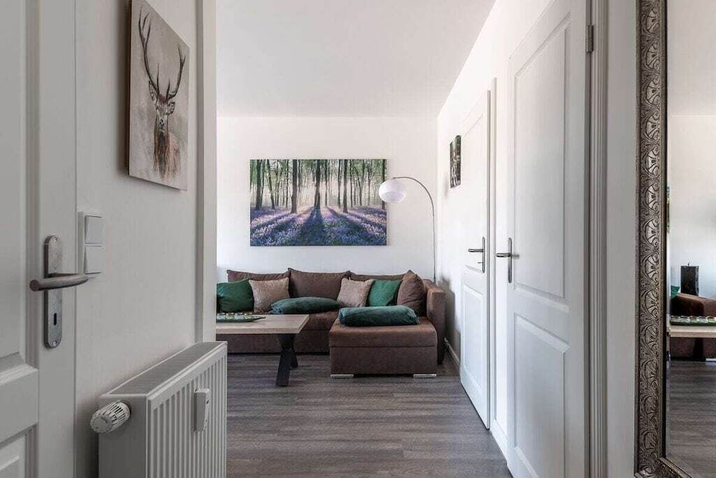 A hallway with a radiator, a sofa with cushions, a painting of a forest, and a door with a decorative frame.
