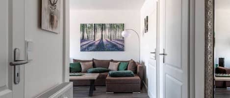 A hallway with a radiator, a sofa with cushions, a painting of a forest, and a door with a decorative frame.